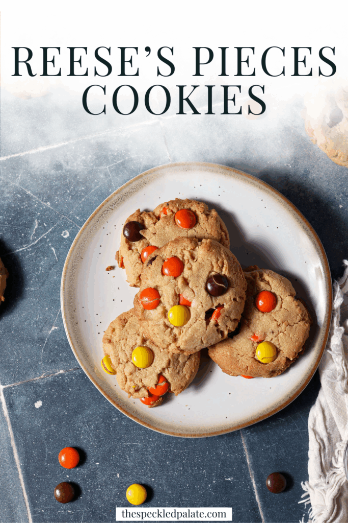 A plate of Reese’s Pieces Peanut Butter Cookies surrounded by more cookies and scattered Reese’s pieces on a dark teal tiled surface next to a white linen with the text reese's pieces cookies