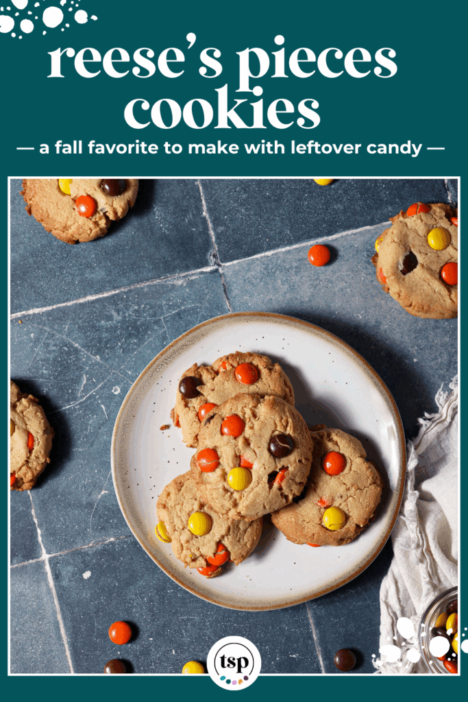 A plate of Reese’s Pieces Peanut Butter Cookies surrounded by more cookies and scattered Reese’s pieces on a dark teal tiled surface next to a white linen with the text reese's pieces cookies a fall favorite to make with leftover candy