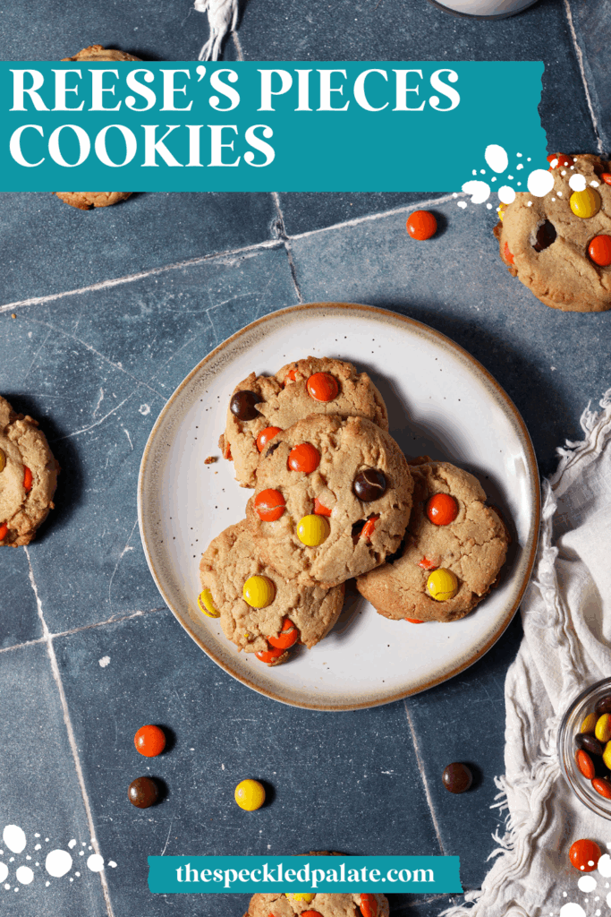 A plate of Reese’s Pieces Peanut Butter Cookies surrounded by more cookies and scattered Reese’s pieces on a dark teal tiled surface next to a white linen with the text reese's pieces cookies