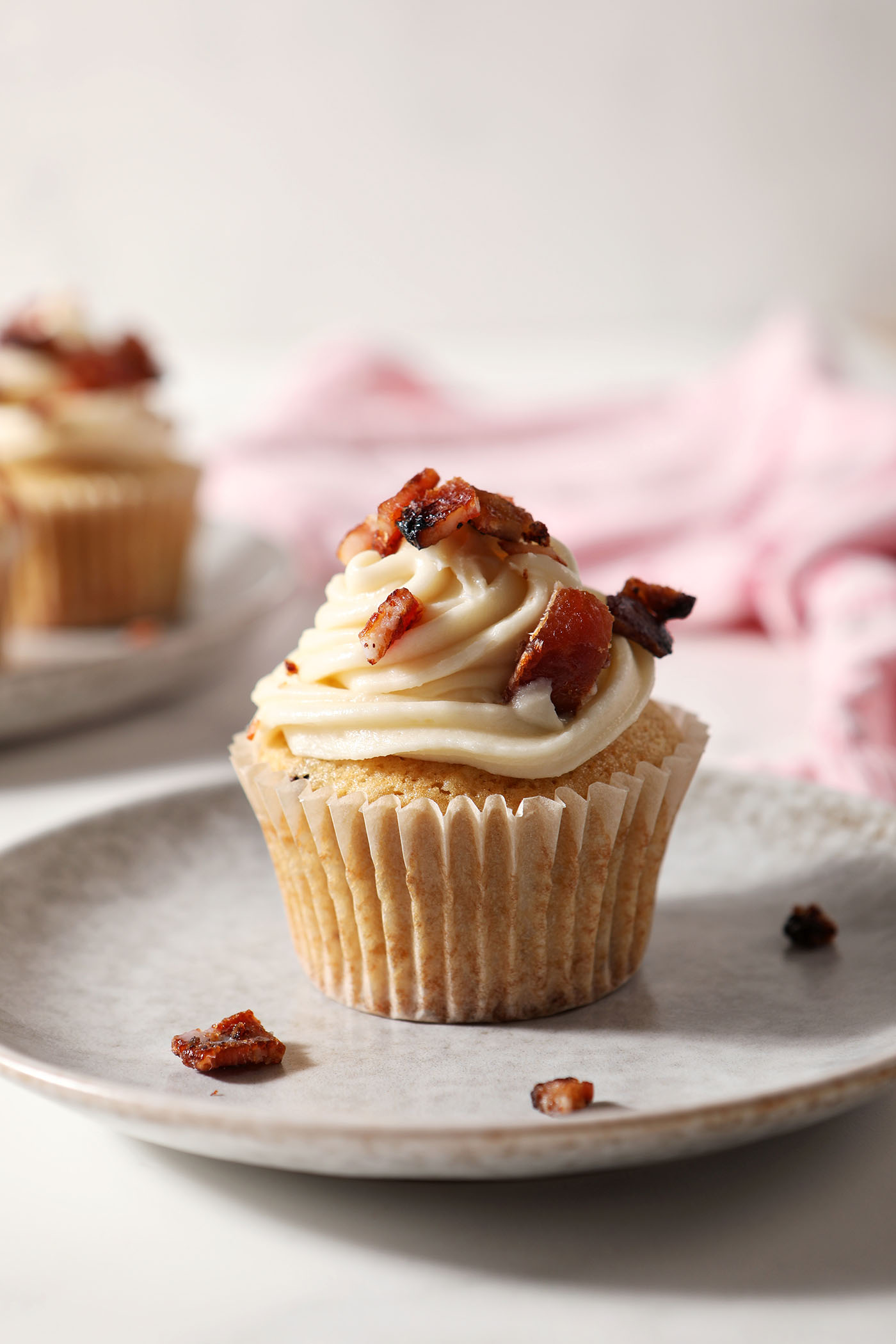 A Bourbon Maple Bacon Cupcake topped with salted maple frosting and chopped maple bacon sits on a grey plate in front of a pink linen
