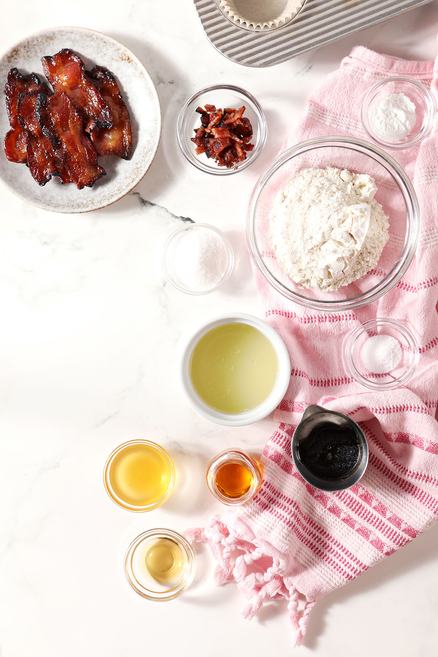 Ingredients to make bourbon maple bacon cupcakes in bowls and on plates on a marble surface with a pink linen