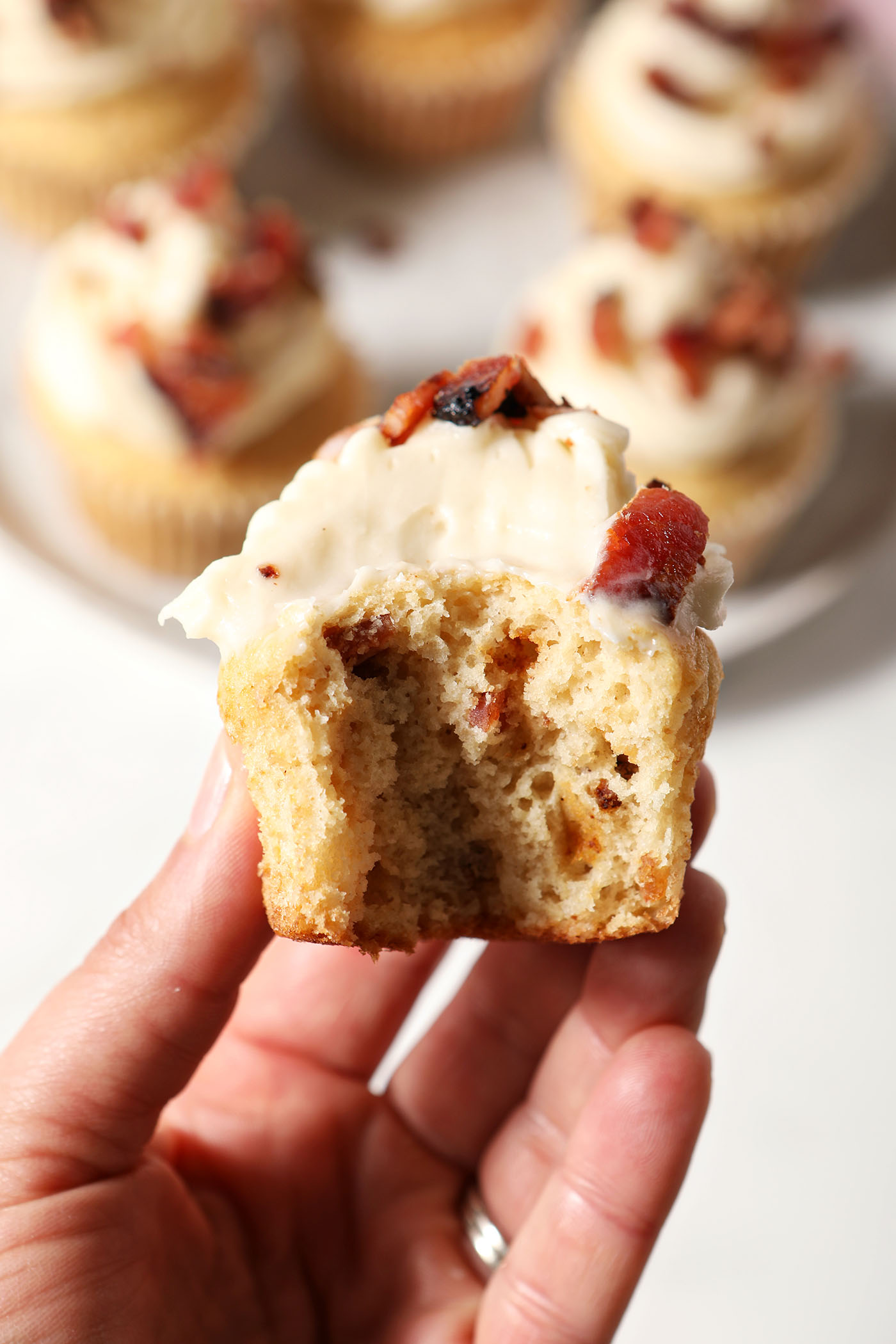 A hand holds a bitten-into maple bacon cupcake with bourbon above a plate of more decorate cupcakes