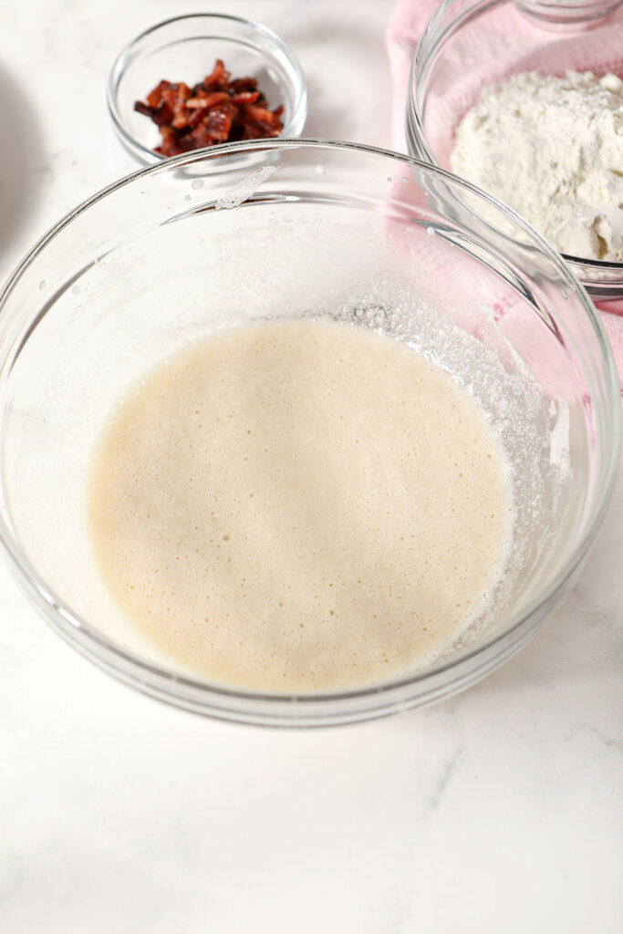 Wet ingredients in a bowl on a marble surface next to a bowl of dry ingredients and a small bowl of chopped bacon