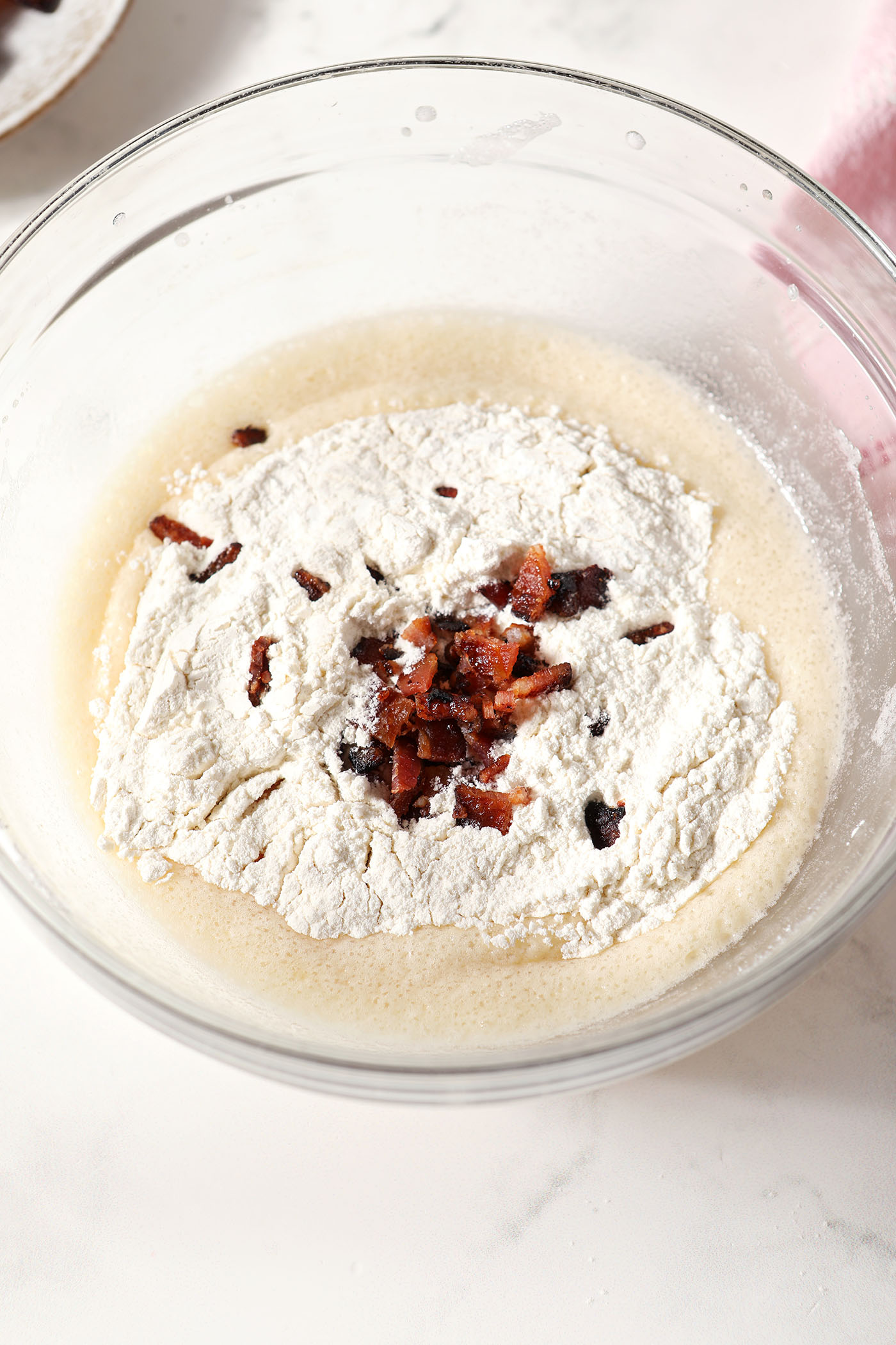 Dry ingredients and homemade bacon bits sit on top of wet ingredients for cupcakes in a large mixing bowl