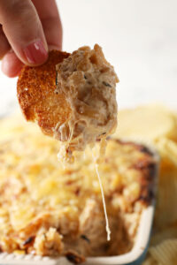 Cheese drips from a toasted crostini after dipping into gooey caramelized onion dip