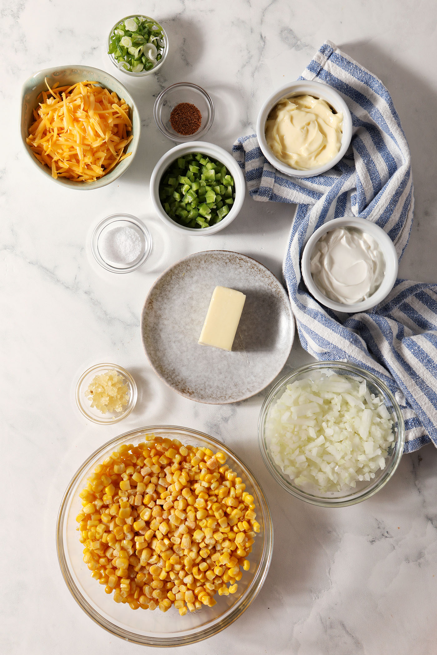 Ingredients to make a creamy corn dip from above in bowls on a marble surface next to a blue and white striped linen