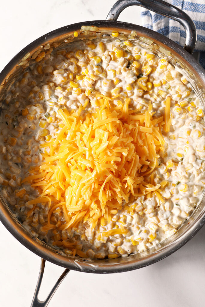 Shredded cheddar cheese on top of creamy corn dip in a skillet, before mixing in