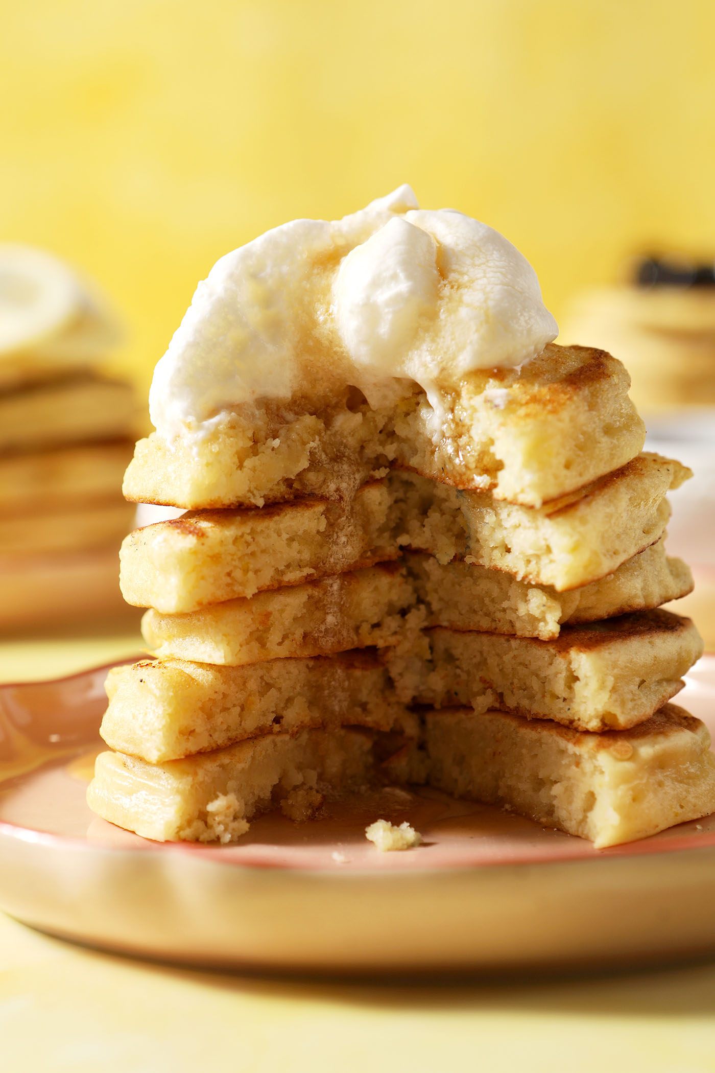 A cut into stack of pancakes topped with whipped cream and maple syrup on a pink plate on a yellow surface