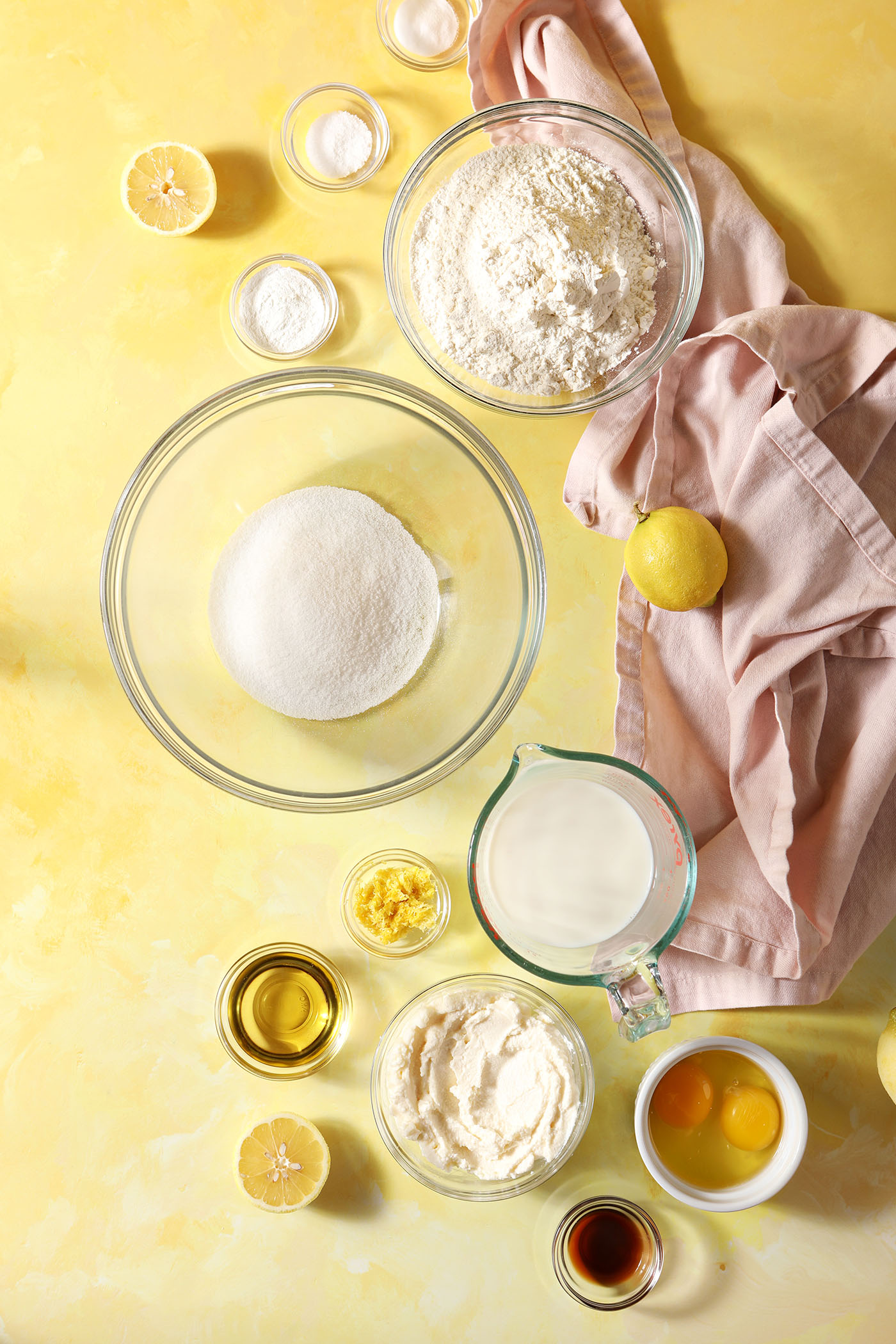 Ingredients to make lemon pancakes with ricotta in bowls and cups on a yellow surface next to a light pink linen