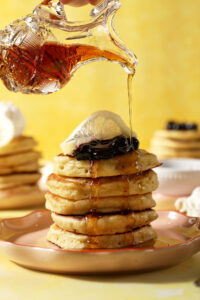 Maple syrup drizzles on top of a stack of lemon pancakes topped with blueberry compote and a lemon twist