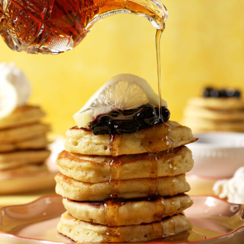 Maple syrup drizzles on top of a stack of lemon pancakes topped with blueberry compote and a lemon twist
