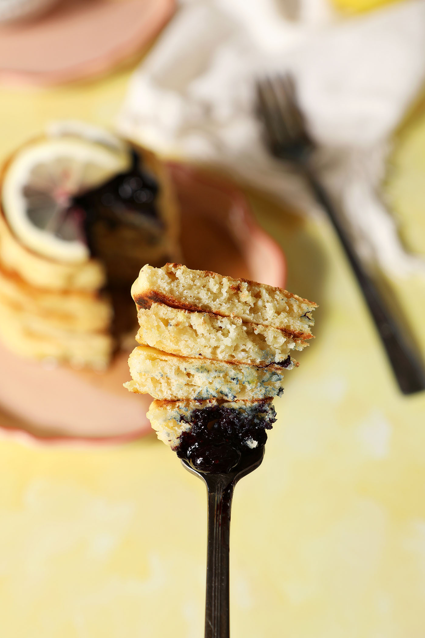 A fork holds a large bite of lemon ricotta pancakes above a pink plate holding a stack on a yellow surface