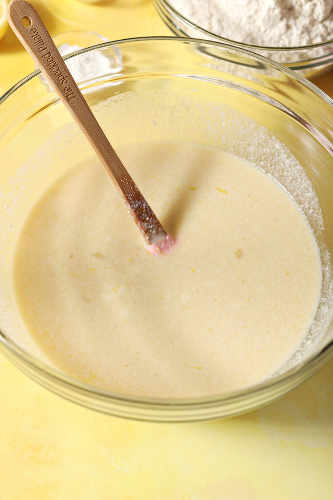 Wet ingredients for pancakes in a large bowl with a rubber spatula on a yellow surface
