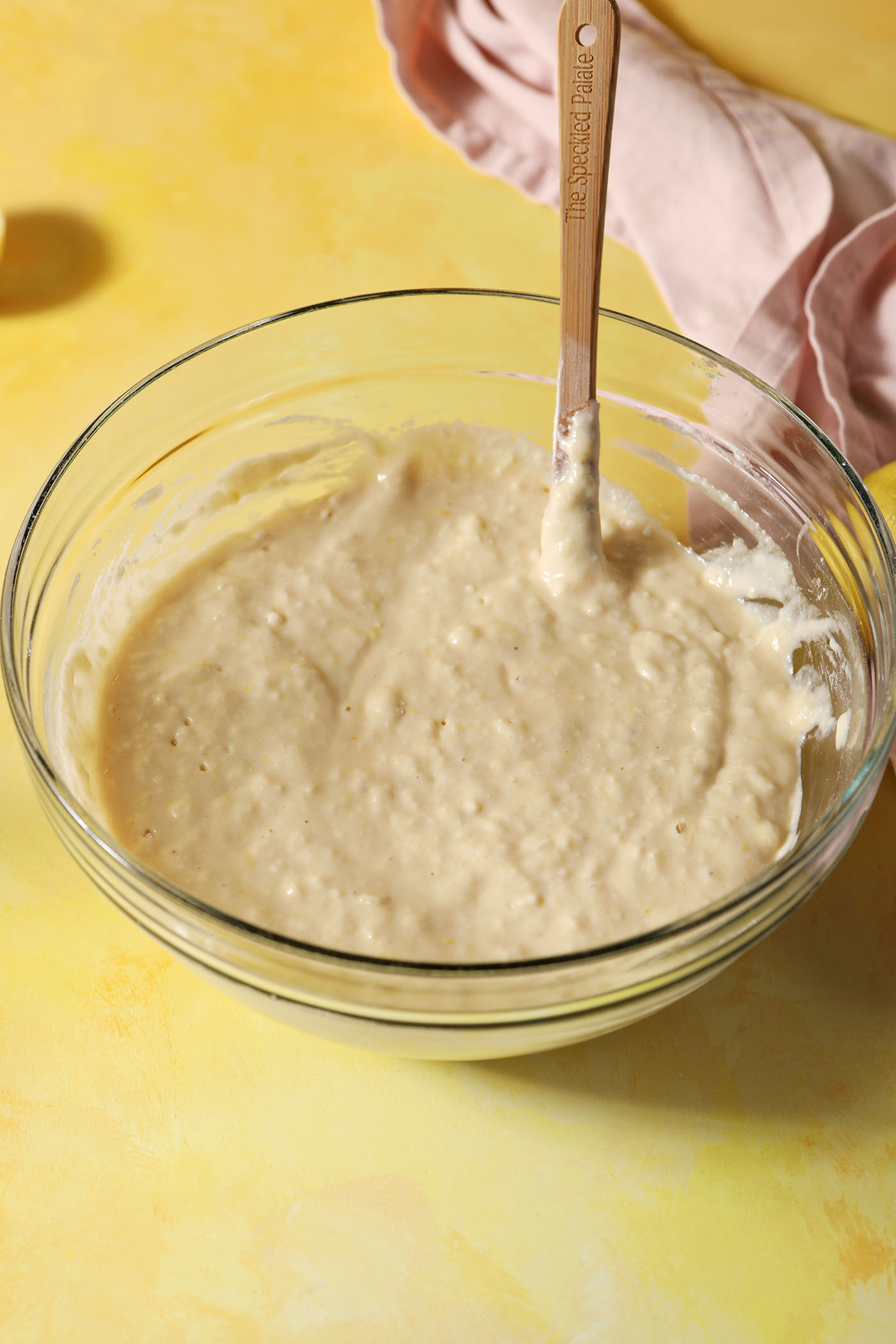 Lemon ricotta pancake batter in a bowl with a spatula on a yellow surface