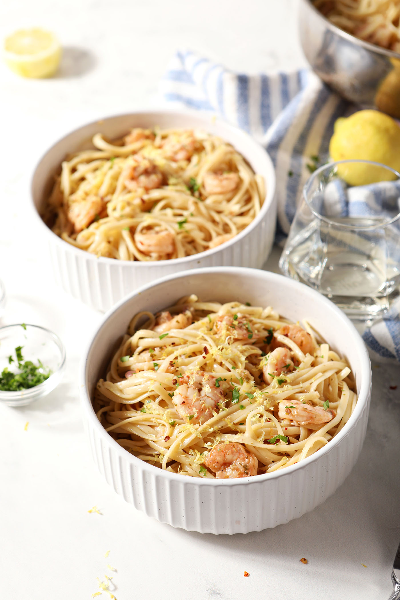 Two bowls holding garlicky lemon shrimp pasta on a white surface next to a blue striped linen, a glass of wine, fresh herbs, lemons and more