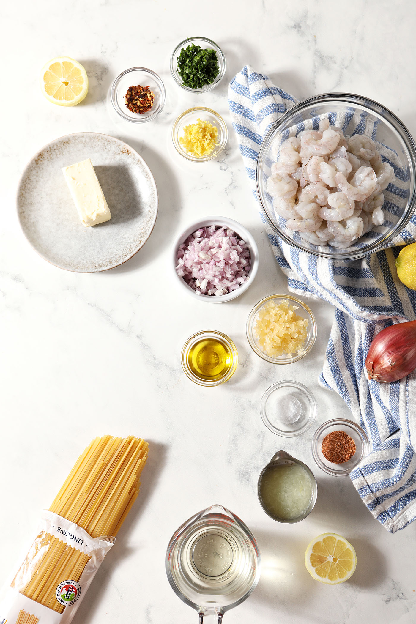 Ingredients to make lemon garlic shrimp pasta in bowls on a marble surface with a blue striped linen