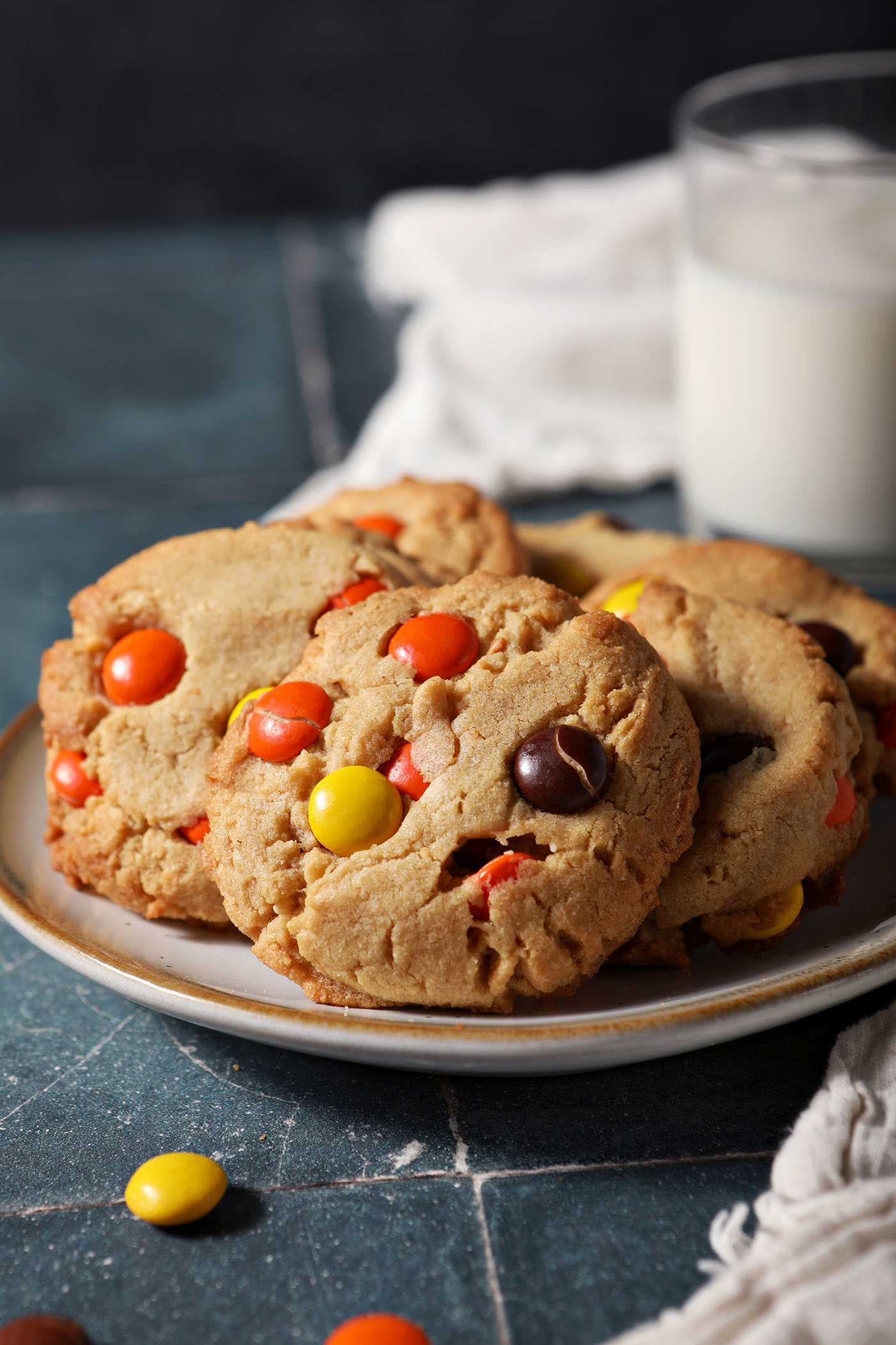 A plate of peanut butter cookies topped with Reese’s pieces sits on a teal tiled surface in front of a glass of milk