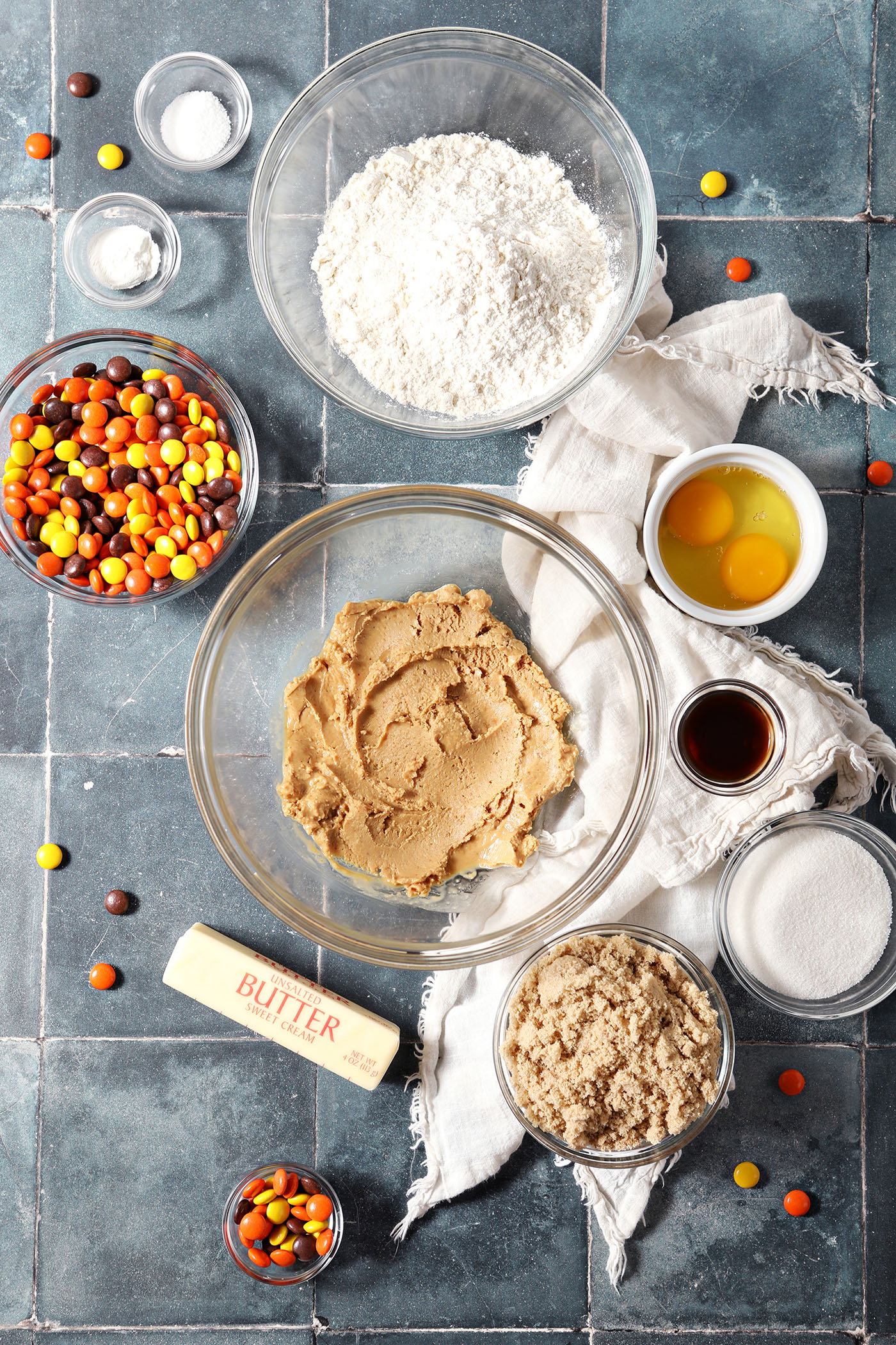 Ingredients to make Peanut Butter Cookies with Reese’s Pieces in bowls on a dark blue tiled surface with a white linen