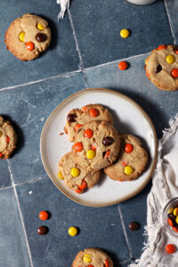A plate of Reese’s Pieces Peanut Butter Cookies surrounded by more cookies and scattered Reese’s pieces on a dark teal tiled surface next to a white linen