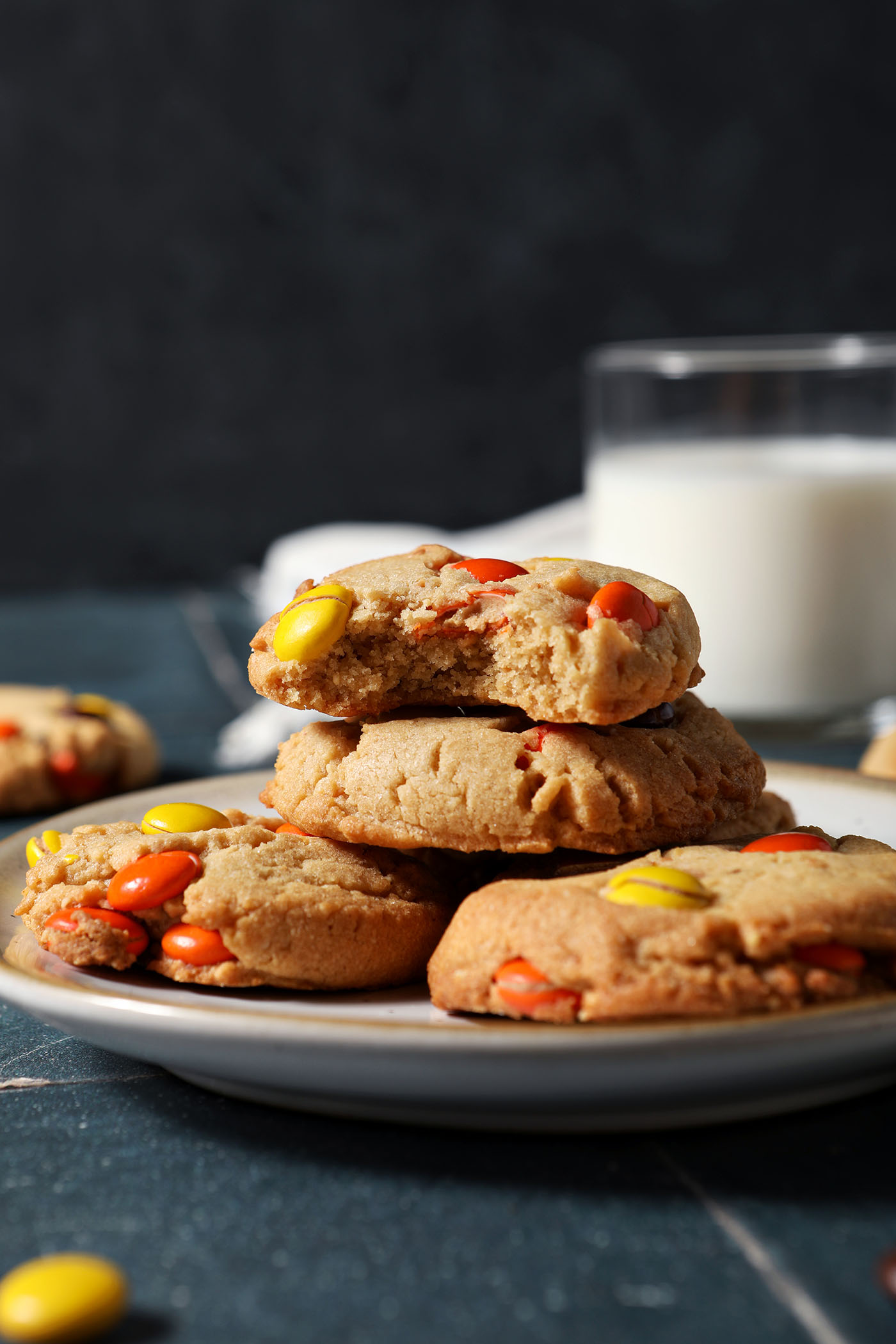 A bitten-into Reese’s Pieces Peanut Butter Cookie sits on top of a stack of cookies on a plate on a dark blue tiled surface