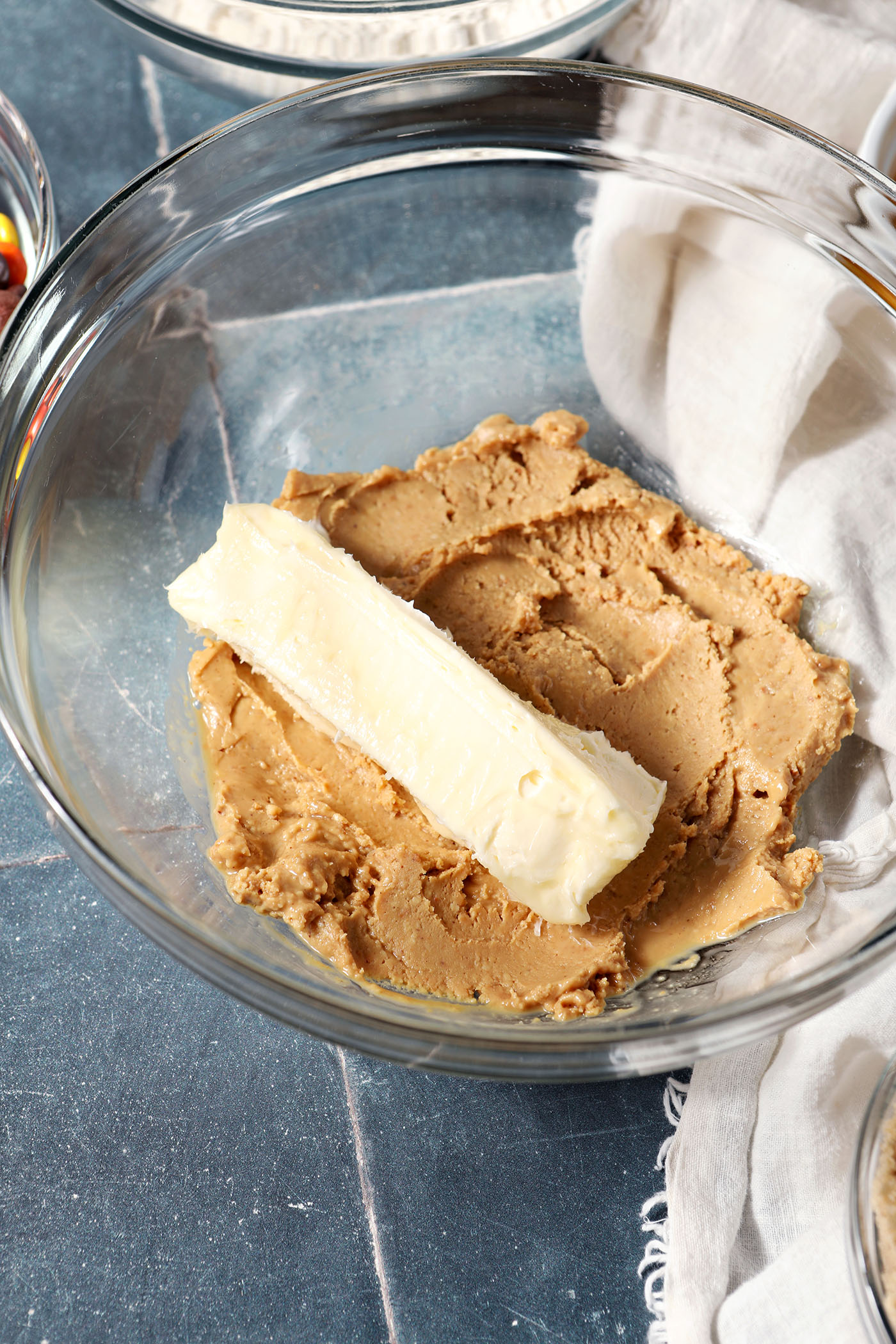 A stick of butter sits on top of peanut butter in a bowl on a dark blue tiled surface