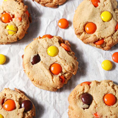 Baked Reese’s Pieces Peanut Butter Cookies surrounded by more Reese’s pieces on parchment paper