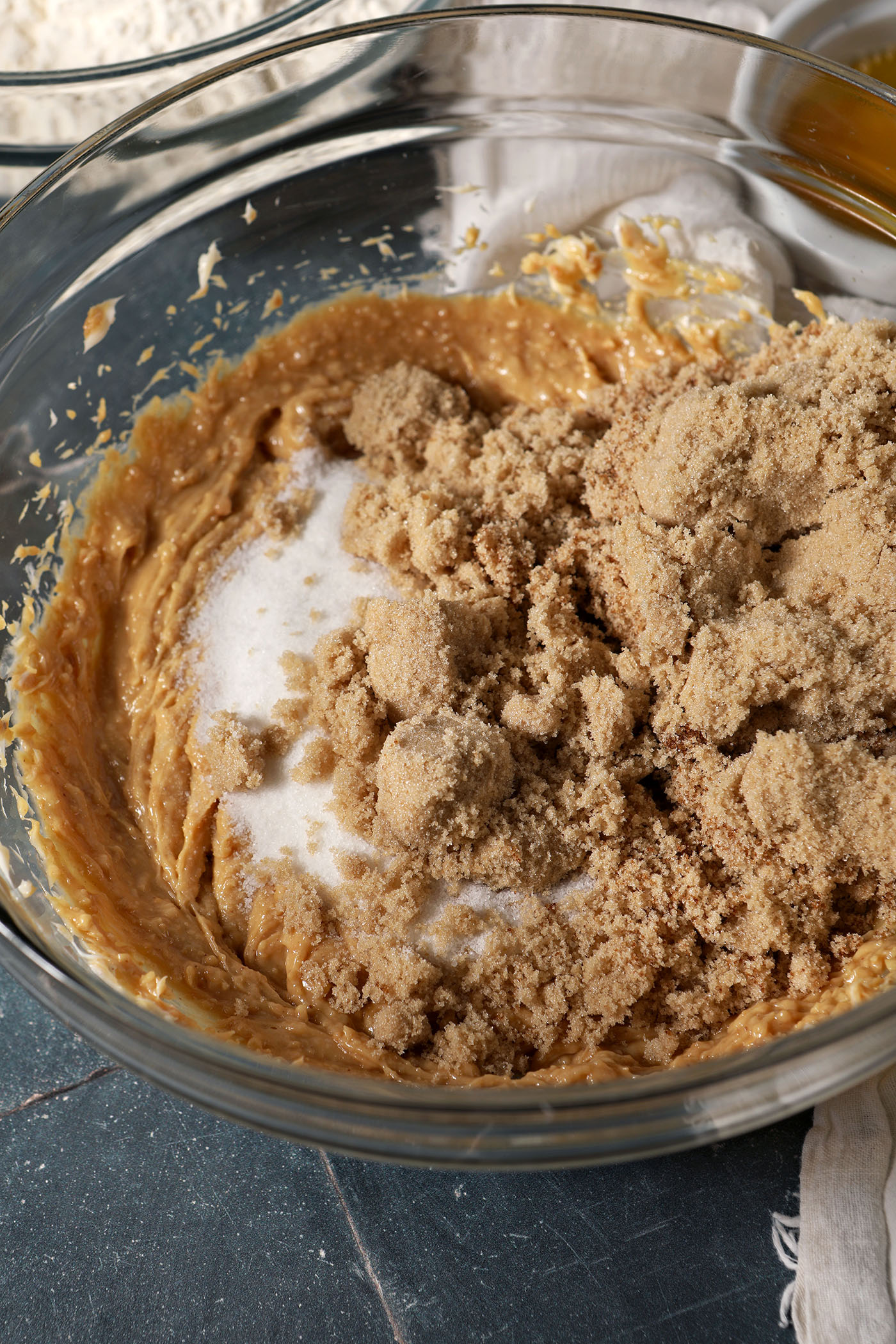 Brown and white sugars on top of whipped peanut butter in a bowl on a dark blue tiled surface