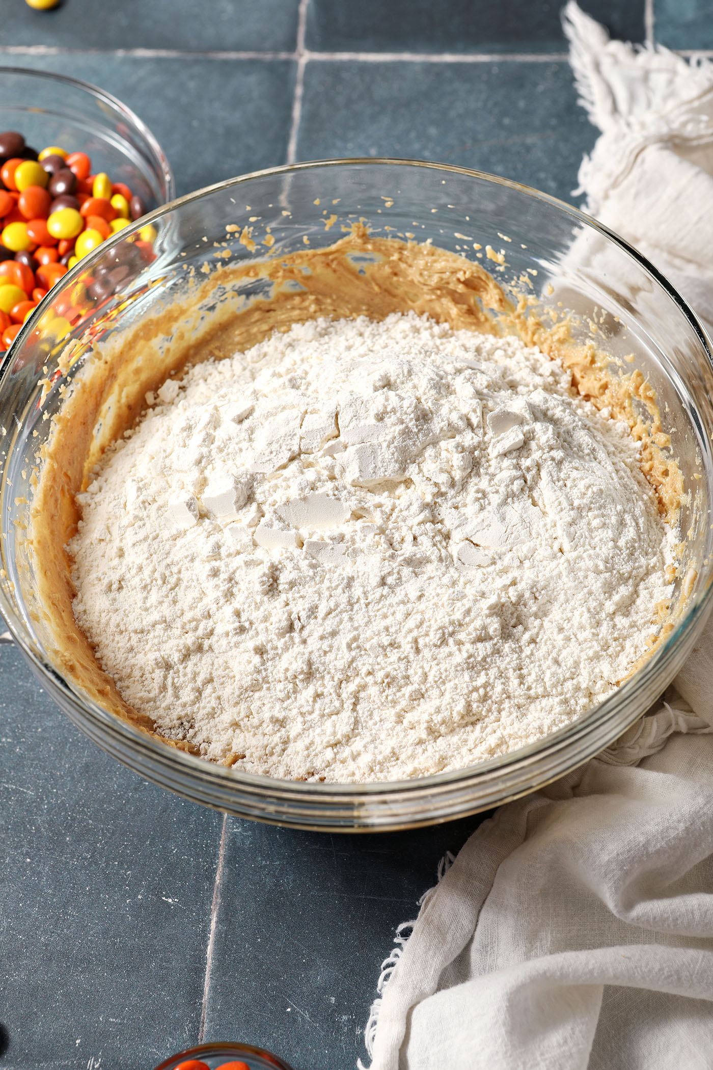 Dry ingredients sit on top of peanut butter cookie wet ingredients in a bowl on a blue tiled surface next to a white linen