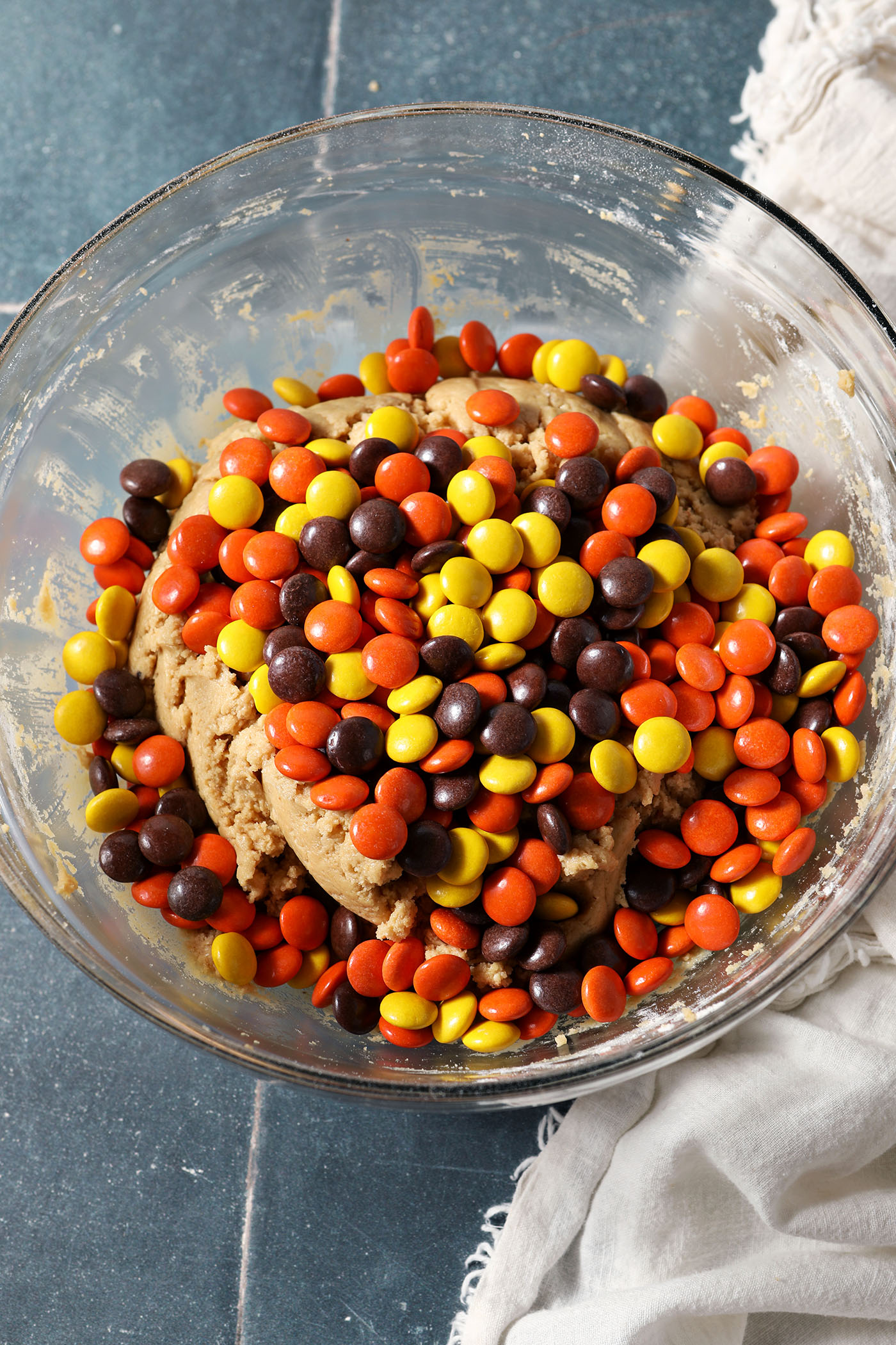 Reese’s pieces sit on top of peanut butter cookie dough on a bowl on top of a blue tiled surface next to a white linen
