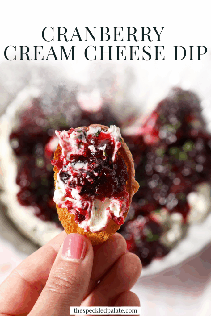 A hand holds a crostini holding cranberry cream cheese dip with jalapeño cream cheese over a platter with the dip with the text cranberry cream cheese dip