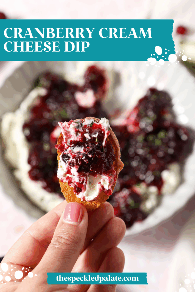 A hand holds a crostini holding cranberry cream cheese dip with jalapeño cream cheese over a platter with the dip with the text cranberry cream cheese dip