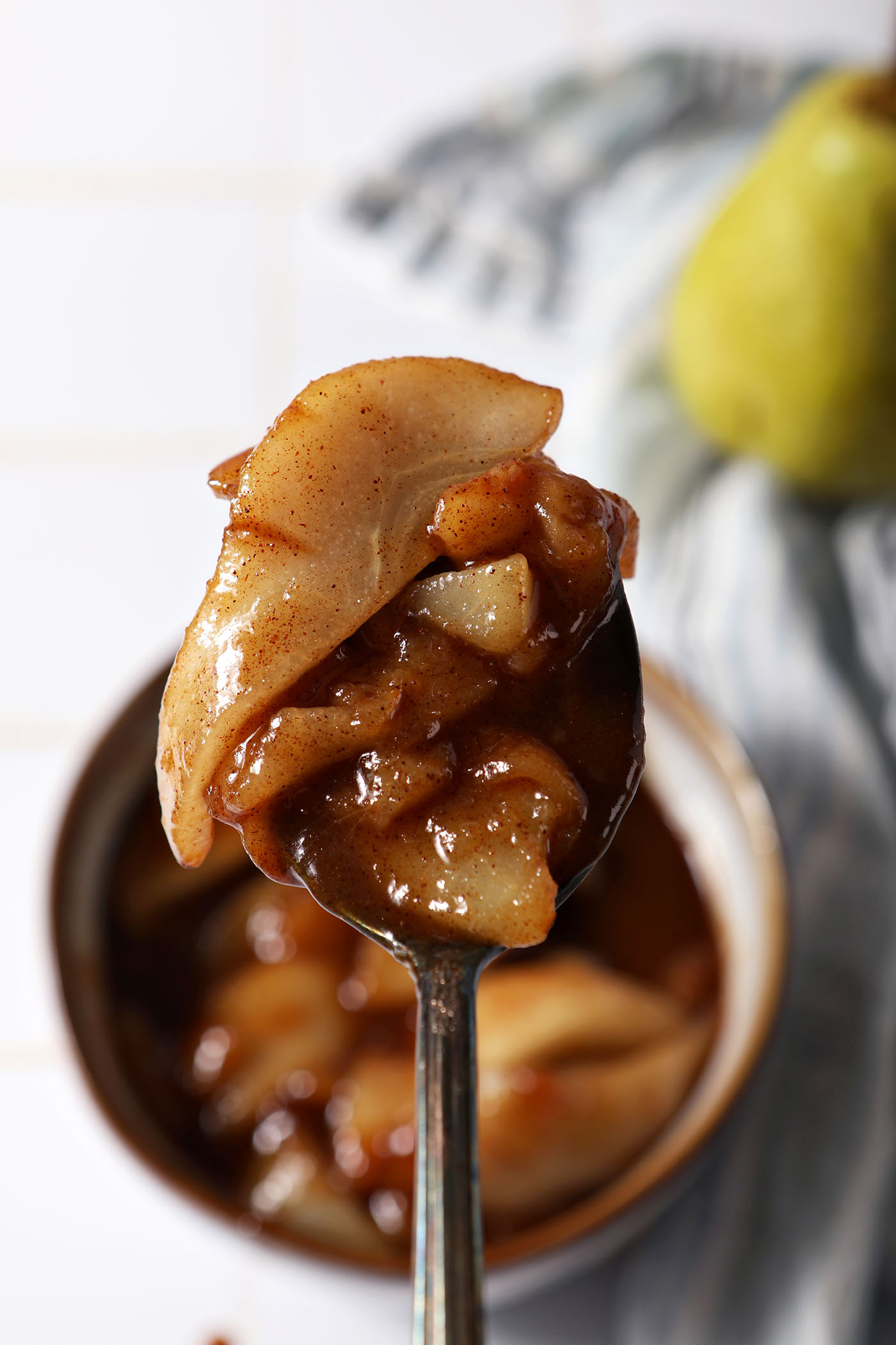 A spoon holds a scoop of caramelized pears above a bowl of more on a tiled surface next to a blue linen and a green pear