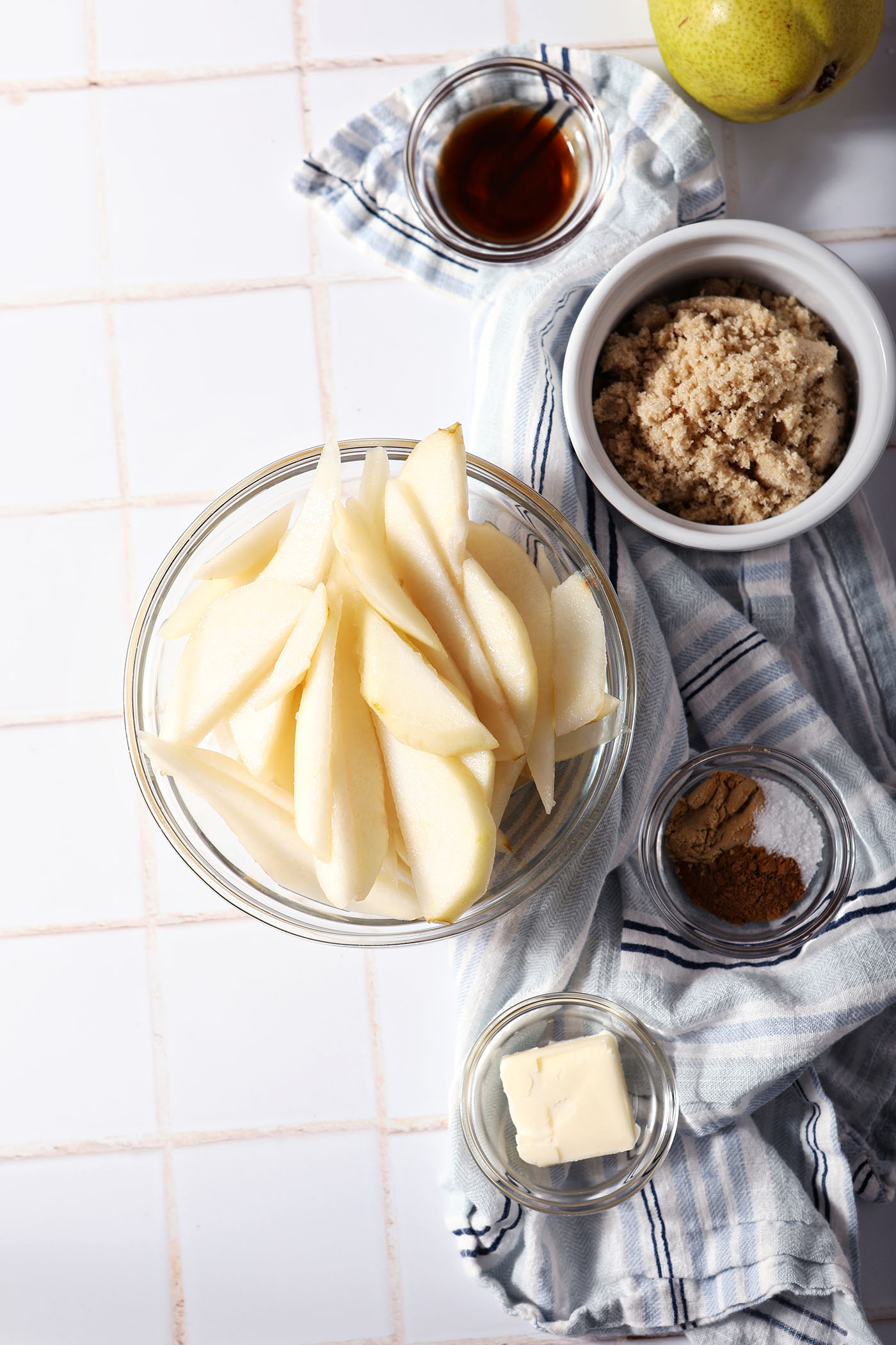 Sliced pears in a bowl next to other ingredients to make caramelized pears on a white tile surface with a blue striped linen