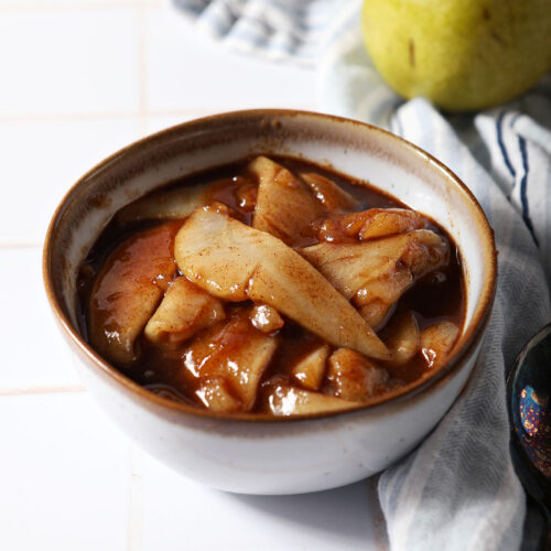 A bowl of caramelized pears on a white tiled surface next to a blue striped linen and a green pear