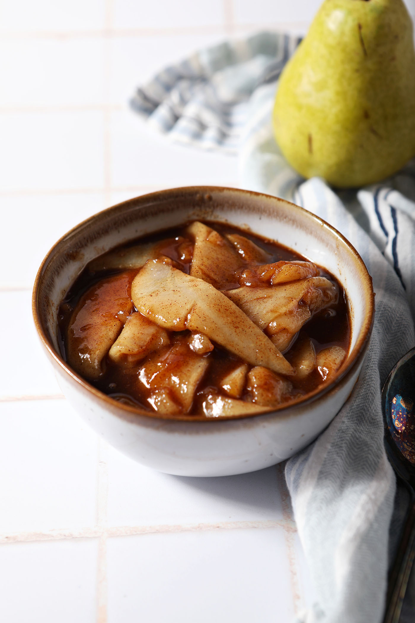 A bowl of caramelized pears on a white tiled surface next to a blue striped linen and a green pear