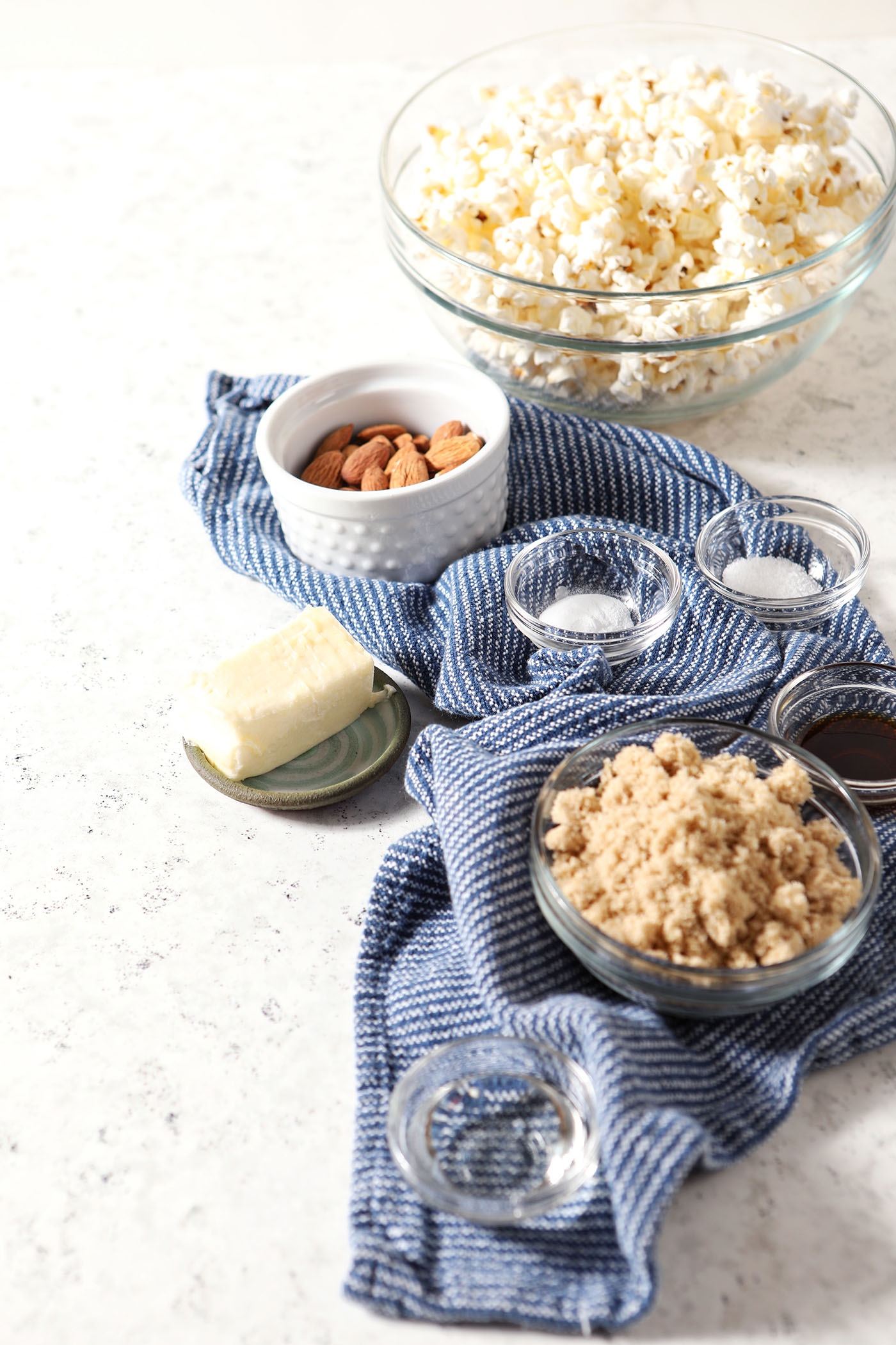 Ingredients to make caramel corn in bowls on a grey tile surface on top of a blue linen