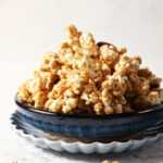 A blue bowl is heaped high with caramel corn on a stone surface, surrounded by caramel corn bits