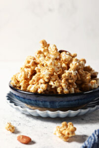 A blue bowl is heaped high with caramel corn on a stone surface, surrounded by caramel corn bits
