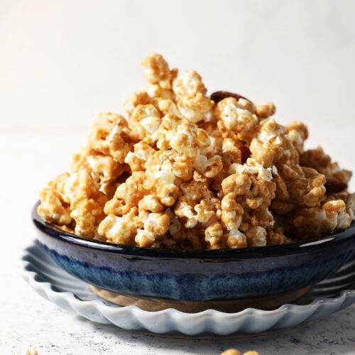 A blue bowl is heaped high with caramel corn on a stone surface, surrounded by caramel corn bits