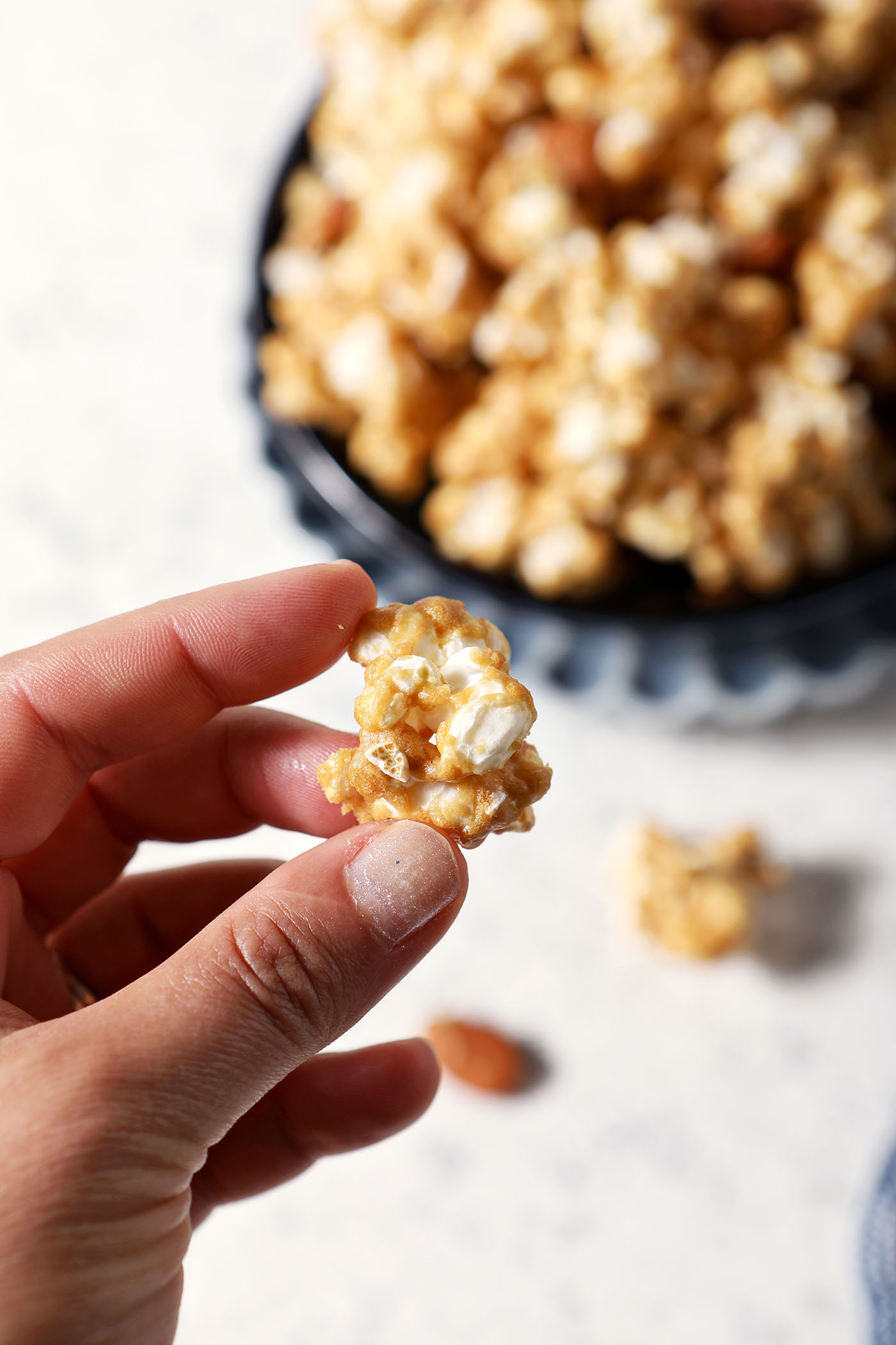 A hand holds a bite of caramel corn above a stone surface and a bowl of more caramel corn
