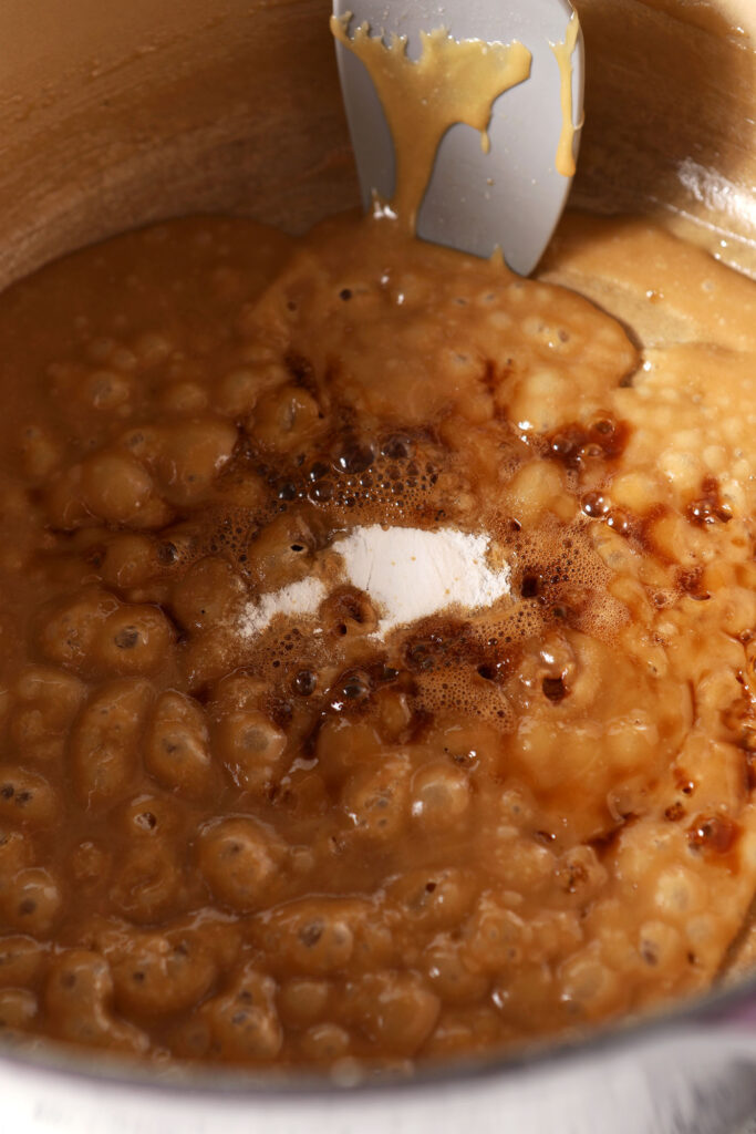 Baking soda and vanilla on top of boiling caramel in a pot next to a grey rubber spatula