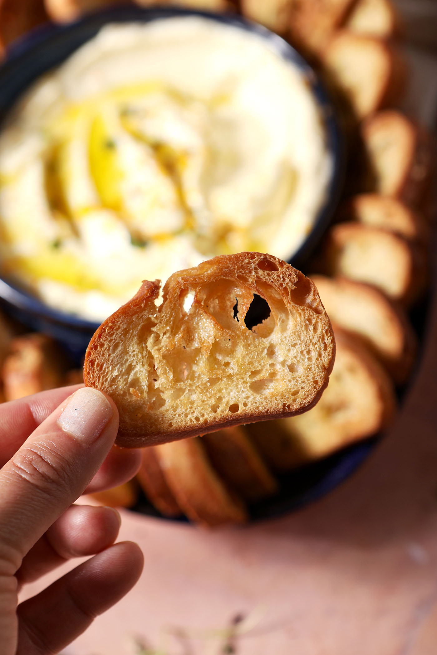 A hand holds a crostini above creamy dip and a platter with more crostini