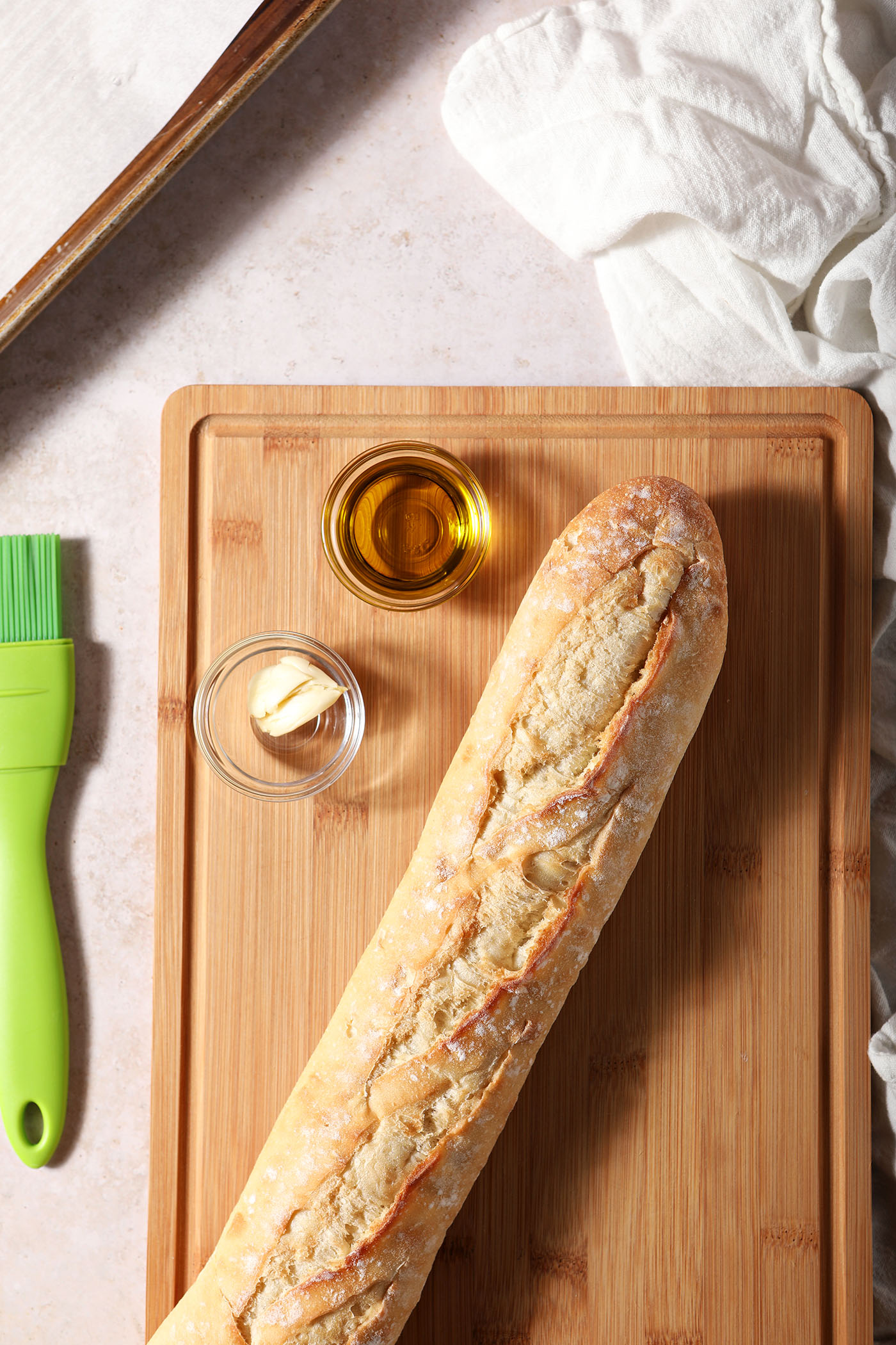 A baguette on a wooden cutting board next to glass bowls of oil and a smashed garlic clive