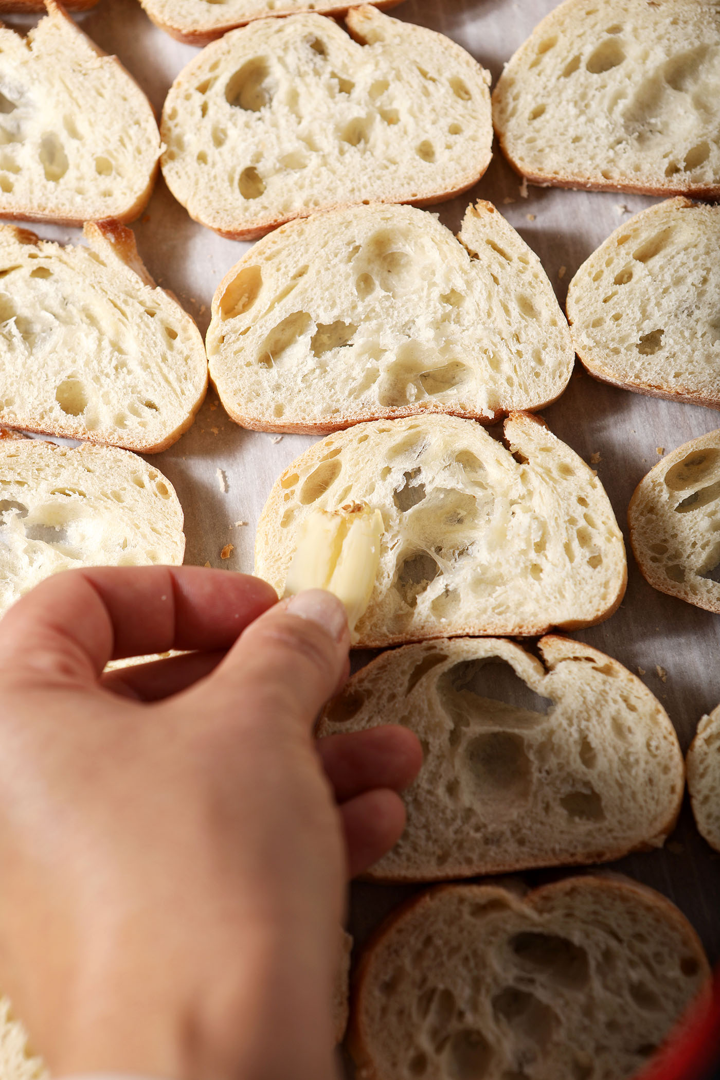 A hand rubs smashed garlic on rounds of baguette on a lined sheet pan