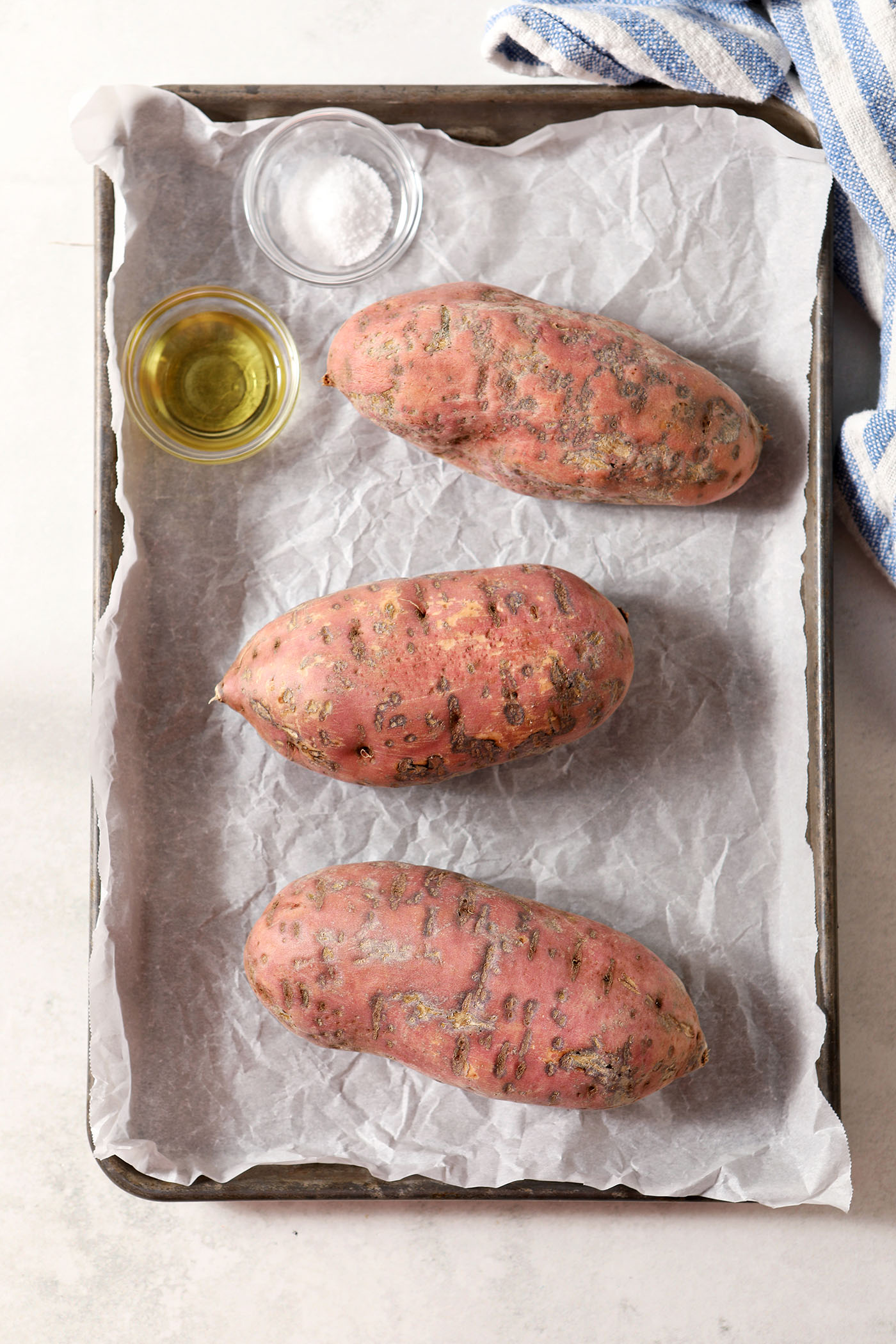 Three sweet potatoes on a sheet pan lined with parchment next to bowls of oil and salt