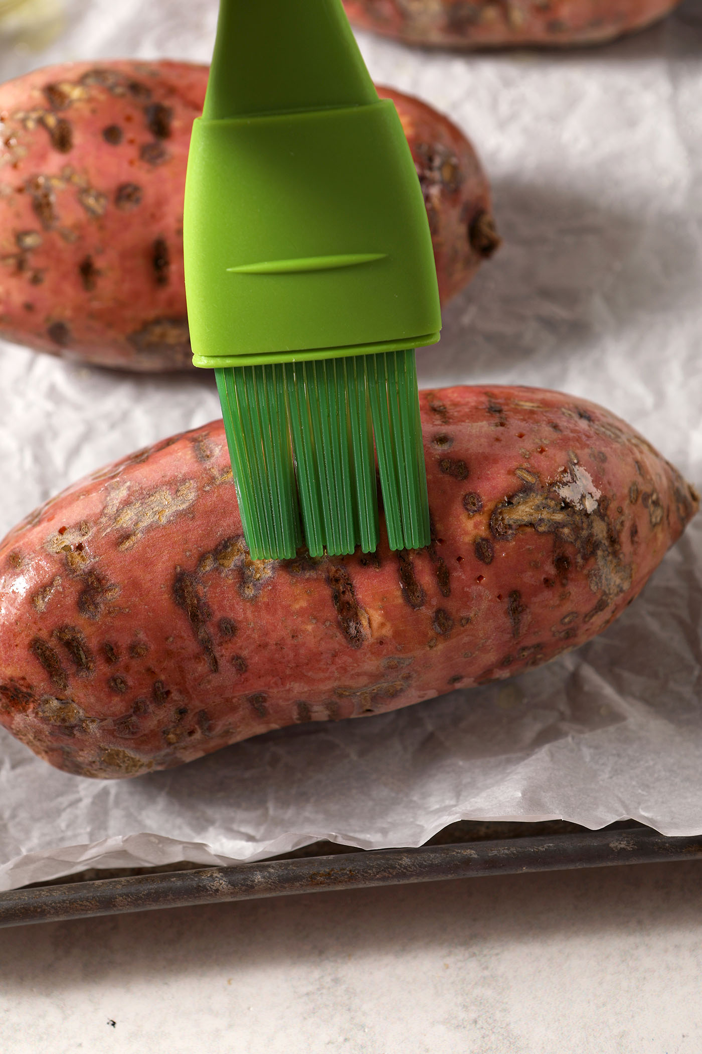A green pastry brush brushes oil on top of a sweet potato on a sheet pan
