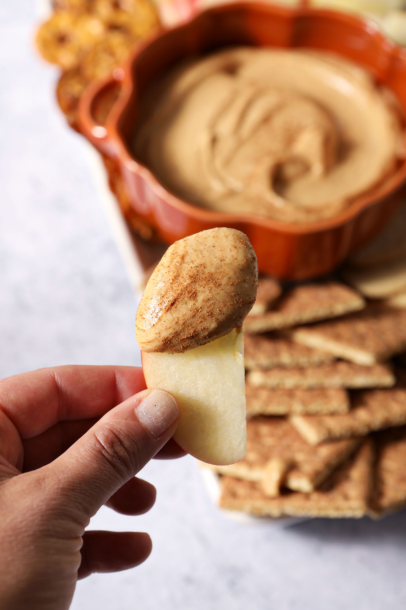A hand holds a slice of apple coated with pumpkin dip above a platter of the dip and dippers