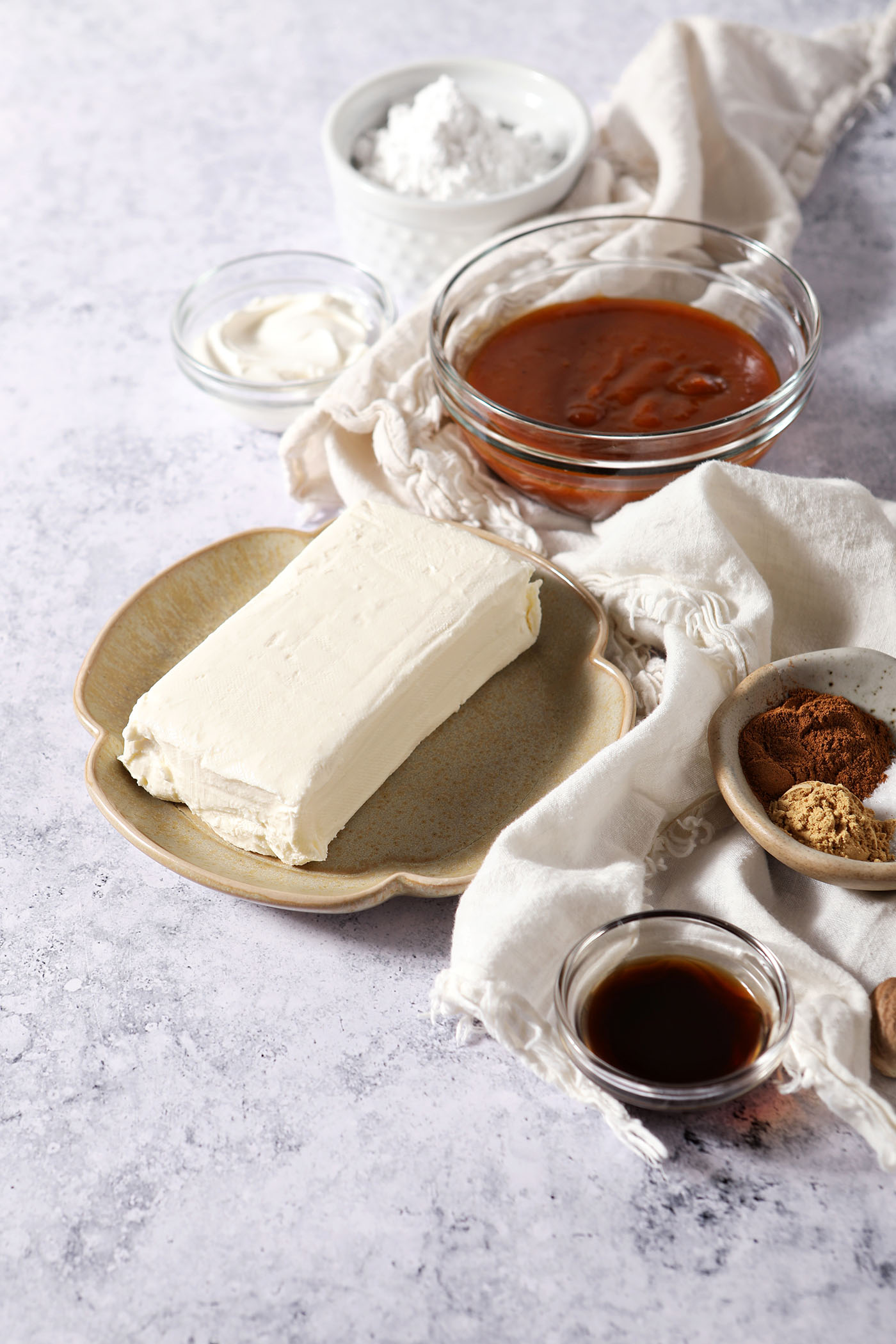 Ingredients to make pumpkin cheesecake dip on plates and in bowls on a grey surface on a white linen