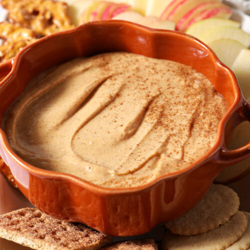 Close up of a pumpkin-shaped bowl holding Pumpkin Cheesecake Dip, surrounded by crackers and apple slices