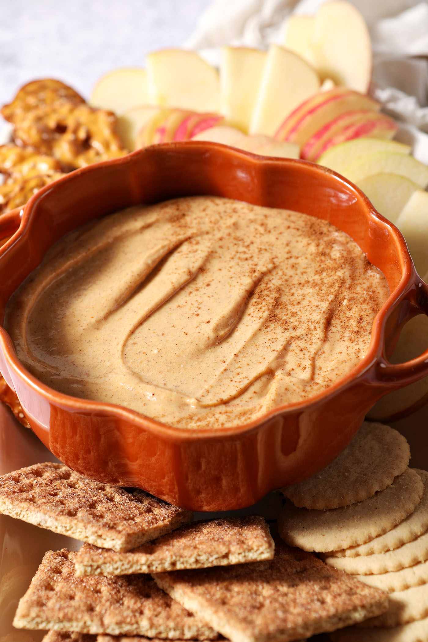 Close up of a pumpkin-shaped bowl holding Pumpkin Cheesecake Dip, surrounded by crackers and apple slices